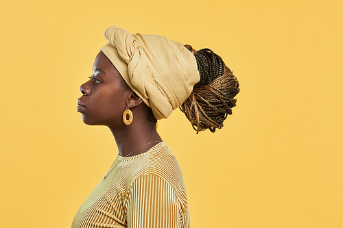 Minimal side view portrait of young African-American woman wearing head wrap and ethnic jewelry while standing against yellow background in studio, copy space