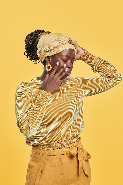 Vertical portrait of trendy African-American woman wearing head wrap and ethnic jewelry while dancing against yellow background in studio