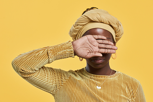 Minimal portrait of young African-American woman wearing head wrap and covering eyes while standing against yellow background in studio, copy space