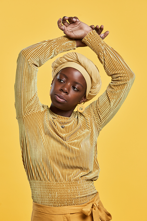 Vertical portrait of confident African-American woman wearing head wrap while dancing against yellow background in studio