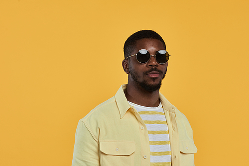 Minimal waist up portrait of trendy African-American man wearing sunglasses and looking at camera while standing against yellow background in studio, copy space