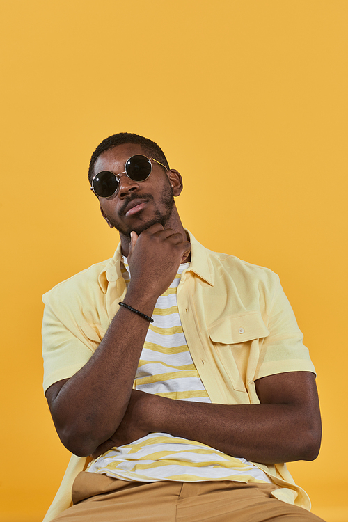 Vertical portrait of handsome African-American man wearing sunglasses and looking at camera while posing against yellow background in studio