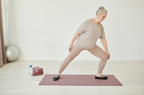 Senior woman in activewear keeping one leg bent in knee while doing physical exercise on mat in home environment