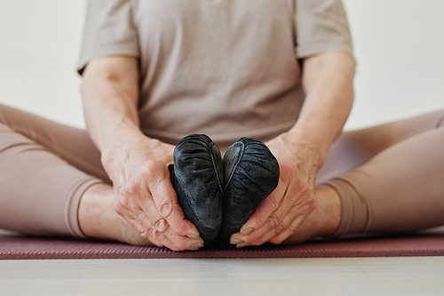 Hands of active senior female holding by her feet in black footwear while sitting on mat and practicing relaxation yoga exercise
