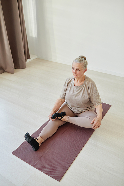 Senior active woman with grey hair sitting on mat and keeping left leg bent in knee on the right one during yoga exercise
