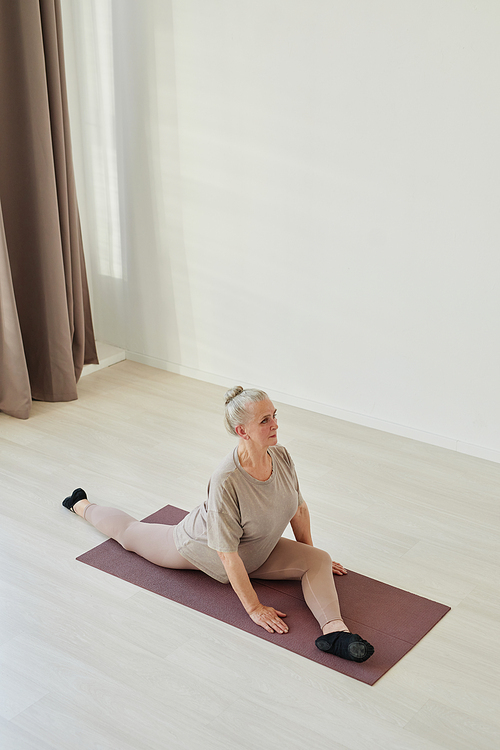 Active senior woman in grey activewear making effort while sitting on mat in yoga position and practicing asanas in living-room