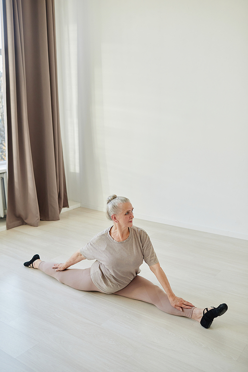 Vertical portrait of aged active woman in sports clothes sitting in yoga position of twine during home workout on the floor