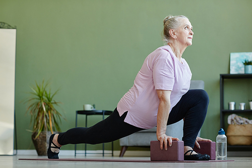 Active senior female with yoga blocks standing with one leg stretcched and another bent in knee while exercising on the floor