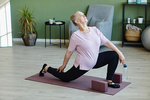 Contemporary senior female in t-shirt and leggins looking upwards during practice of yoga exercise on mat in living-room