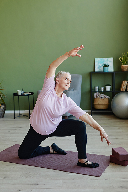 Active senior female in black leggins and beige t-shirt doing physical exercise on yoga mat against domestic plants and furniture in living-room