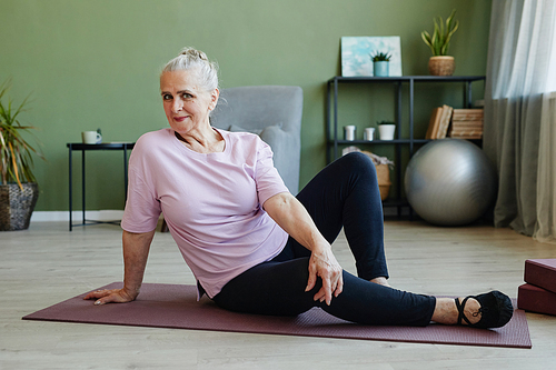 Happy active senior female in sports clothes looking at camera while sitting on yoga mat and exercising in living-room at leisure