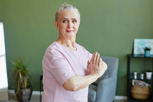 Senior woman with grey hair keeping her hands put together against chest and looking at camera while practicing yoga