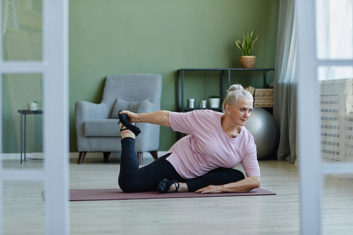 Active senior female practicing yoga asanas on the floor of living-room while sitting on mat and stretching legs and arms