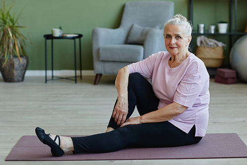 Happy senior female in sports clothes sitting on yoga mat on the floor of living-room on background of armchair and small table