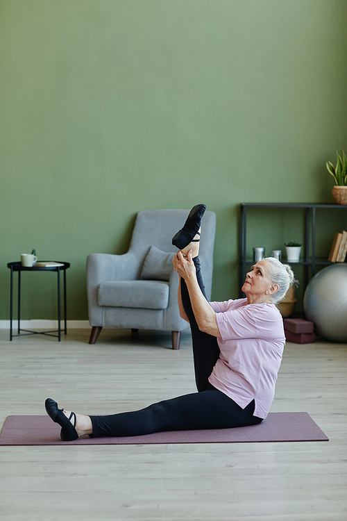 Senior sportswoman in beige t-shirt and black leggins holding and raising stretched leg while exercising on yoga mat