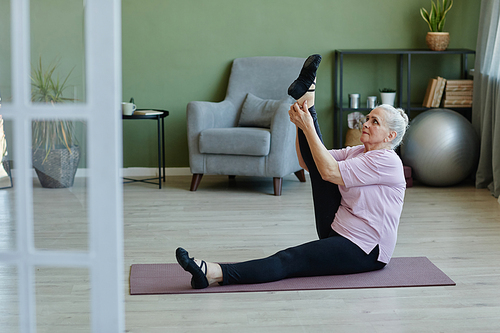 Senior woman in activewear keeping right leg raised and stretched while doing physical exercise on yoga mat in living-room