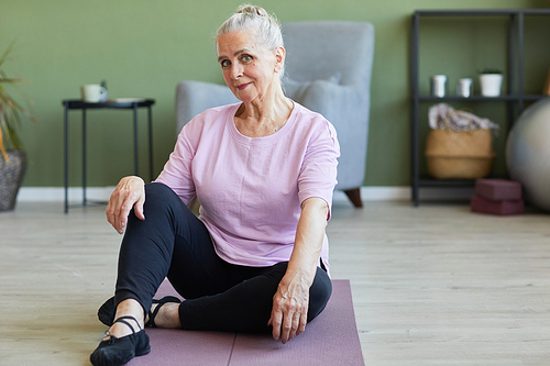 Happy active senior female in sports clothes looking at camera while sitting on yoga mat in living-room on background of furniture