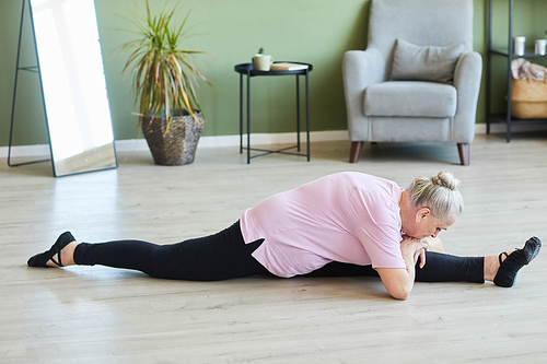 Flexible senior female in black leggins and beige t-shirt sitting on twine on the floor of living-room with her legs split and stretched