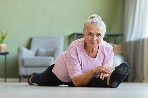 Senior active woman in sports clothes looking at camera while sitting in yoga position called hanumanasana on the floor of living-room
