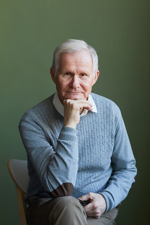Serious elderly man in light blue pullover sitting on chair in front of camera against dark green wall and keeping hand by chin