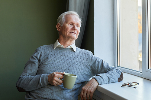 Modern senior man in blue pullover sitting by windowsill in the kitchen, having tea and looking through window