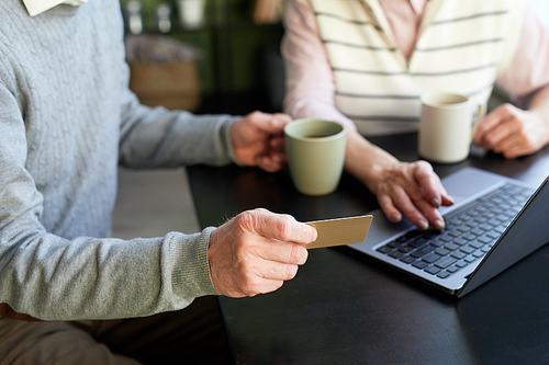 Hand of senior man in blue pullover holding credit card over table while shopping online and ordering takeaway food with his wife