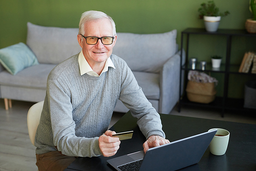 Smiling senior man with credit card looking at camera while sitting by table in front of laptop and shopping online at home