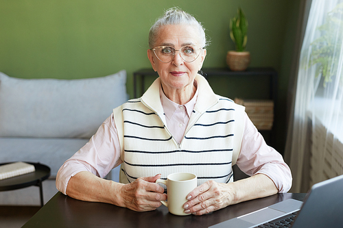 Happy mature female in eyeglasses and casual clothes having tea while sitting by table in front of laptop in living-room
