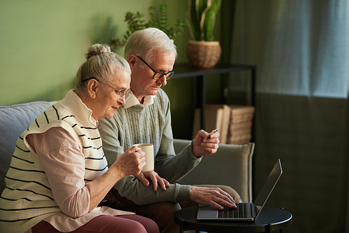 Contemporary senior woman with drink sitting by her husband with credit card paying for purchases during online shopping