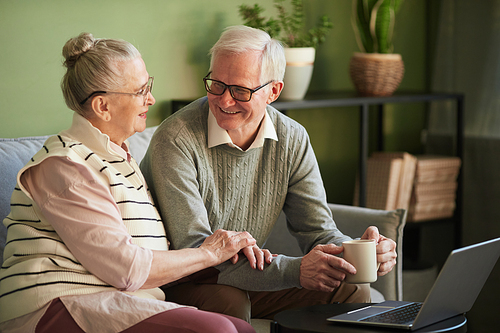 Happy retired husband and wife in casual clothes having talk while sitting on couch in front of laptop in living-room