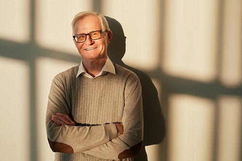 Happy senior man with in beige casual clothes crossing arms by chest while standing by sunlit wall and looking at camera