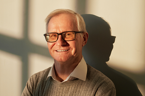 Cheerful aged man in eyeglasses and casual pullover standing by wall inside sunlit room or office and looking at camera