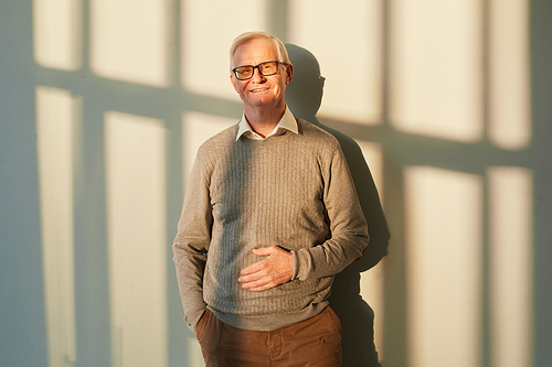 Smiling elderly man in brown pants and pullover and eyeglasses standing against sunlit wall with shadows and looking at camera