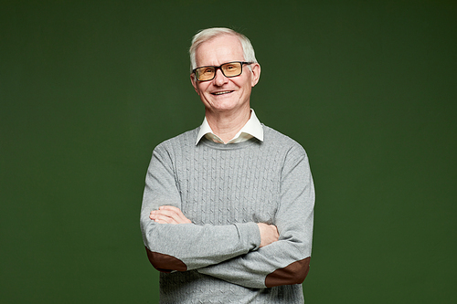 Happy senior male in light blue pullover keeping his arms crossed by chest while standing on dark green background and looking at camera