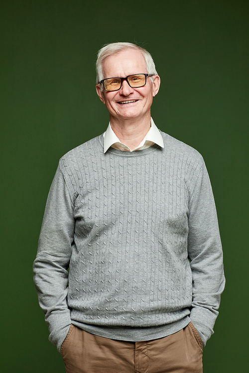 Cheerful elderly man in pants, pullover and eyeglasses standing on dark green background in front of camera