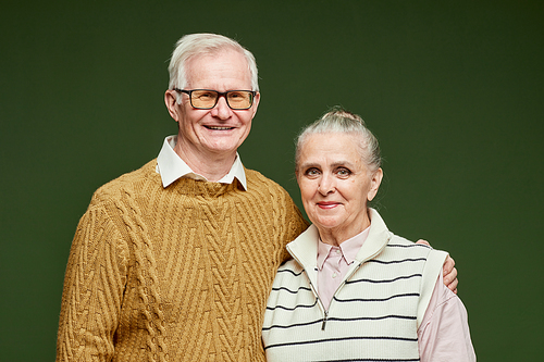 Portrait of happy senior husband in brown pullover embracing his wife in striped casual clothes on dark green background