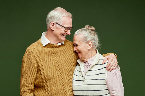 Laughing senior couple in casual clothes standing on dark green background, man in pullover embracing his wife