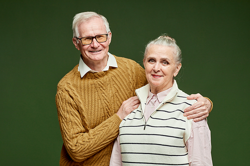 Contemporary cheerful senior couple in pullovers standing on dark green background and looking at camera with smiles