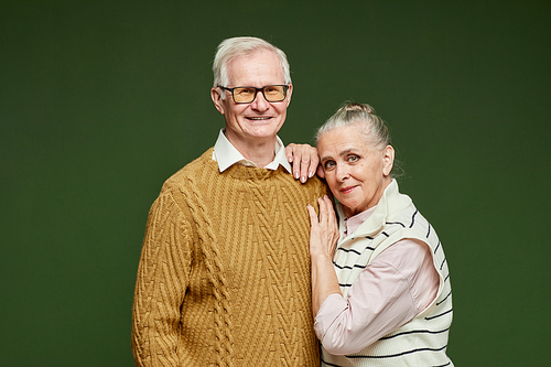 Affectionate elderly husband and wife with grey hair standing against dark green background and looking at camera