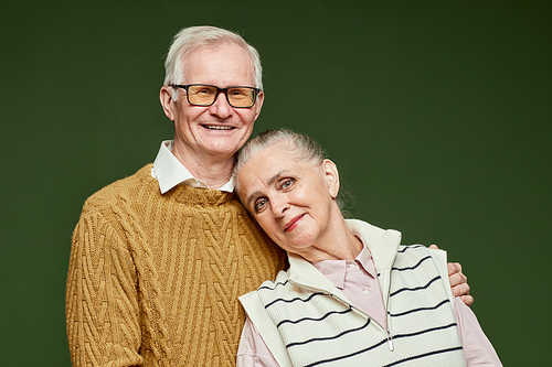 Happy elderly husband and wife in pullovers standing close to one another against dark green background in front of camera