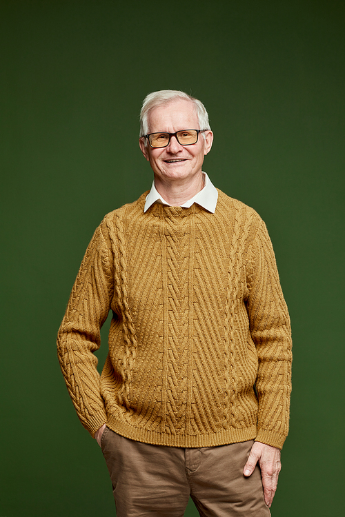 Fashion portrait of cheerful senior man in brown knitted pullover and pants standing in front of camera on dark green background