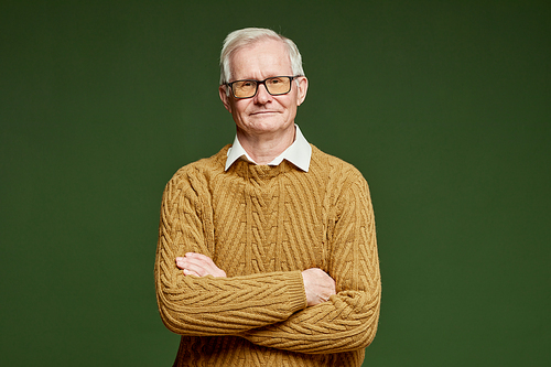 Stylish senior man in knitted sweater of brown color keeping his arms crossed by chest while standing on dark green background