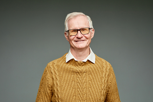 Cheerful aged grey haired man wearing brown knitted sweater and eyeglasses standing against grey color background