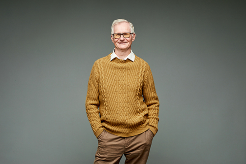 Happy elderly man in knitted sweater and brown pants standing against grey background and looking at camera with smile