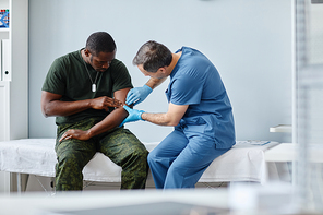 Professional traumatologist wearing protective gloves palpating injured arm of young African American soldier