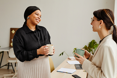 Minimal portrait of two successful young women chatting at coffee break in office, focus on black businesswoman smiling
