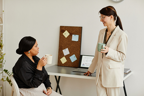Minimal portrait of two successful young women talking at coffee break in office and holding mugs