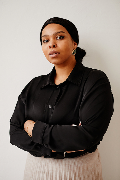 Vertical portrait of successful black businesswoman looking at camera while standing with arms crossed against neutral background