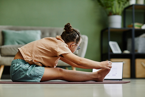 Side view portrait of teenage girl doing stretching exercises while enjoying yoga at home via online lesson