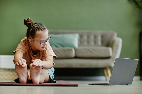 Front view of cute girl with down syndrome doing stretching exercises while enjoying yoga at home via online lesson